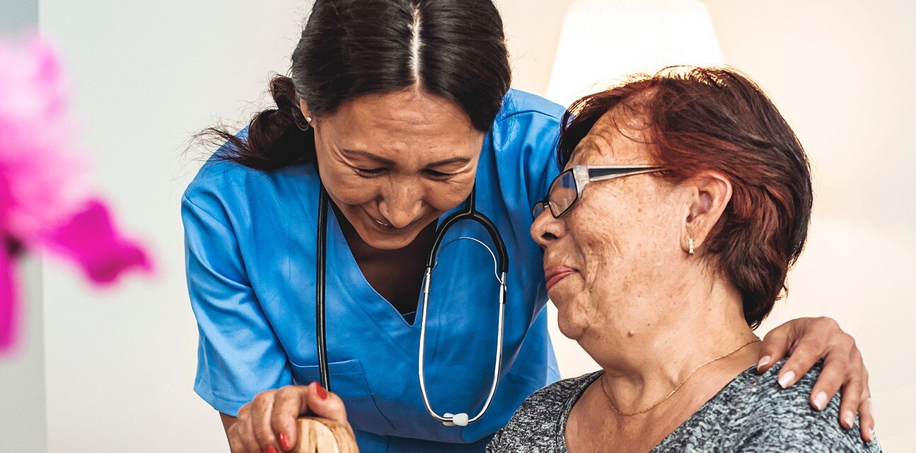 Female nurse in blue fatigues leaning over and holding hand of elderly woman who is smiling back at hear