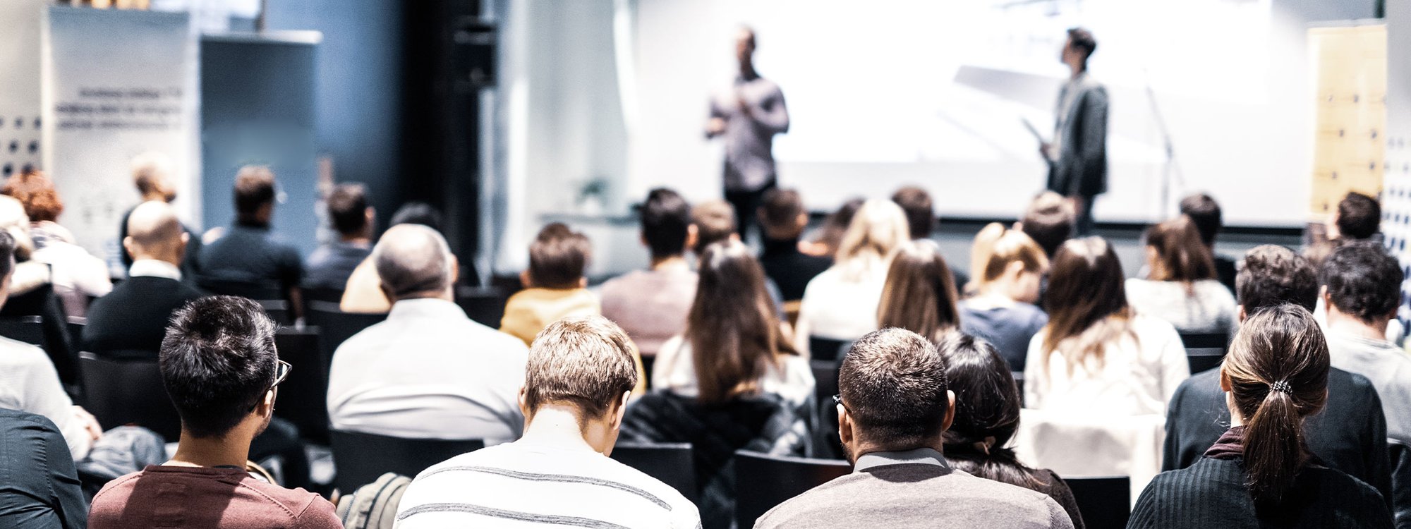 A seated crowd at a trade show watches a presentation