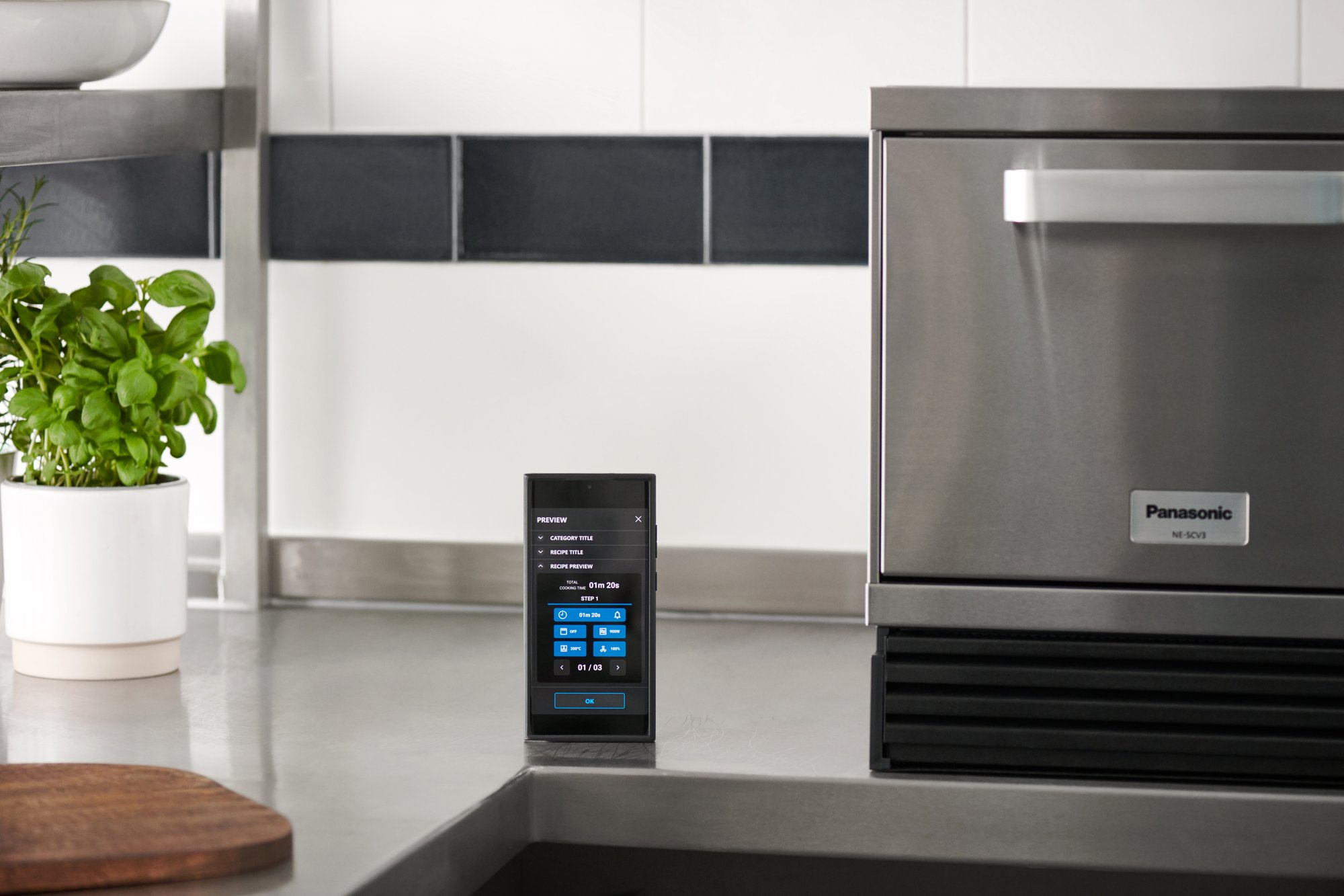 Panasonic NE-SCV3N high speed oven with mobile APP landing page displayed on a smart phone next to it as well as a green plant. 