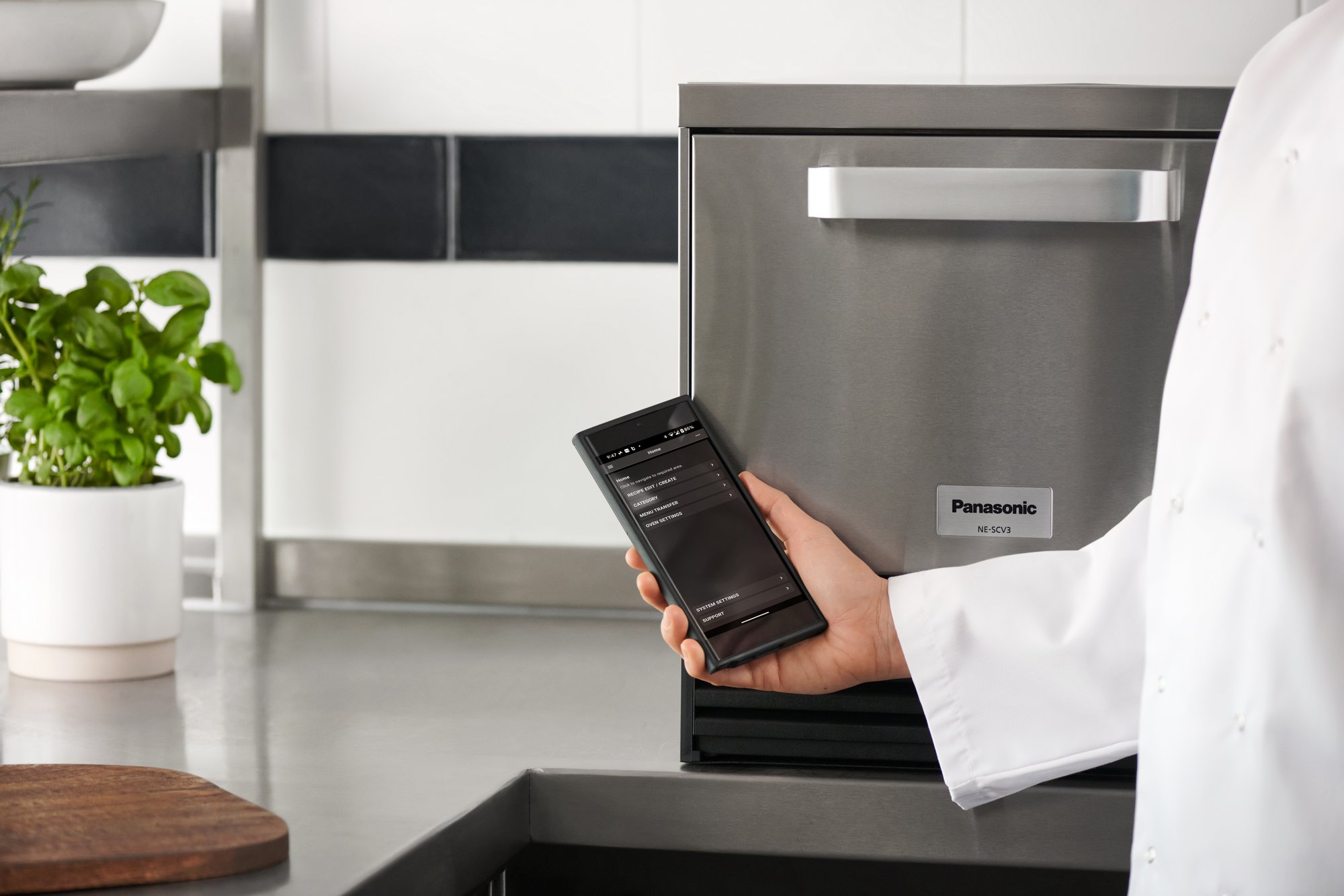A smartphone displaying the SCV Control Hub app is held in front of the Panasonic NE-SCV3N oven in a professional kitchen setting.