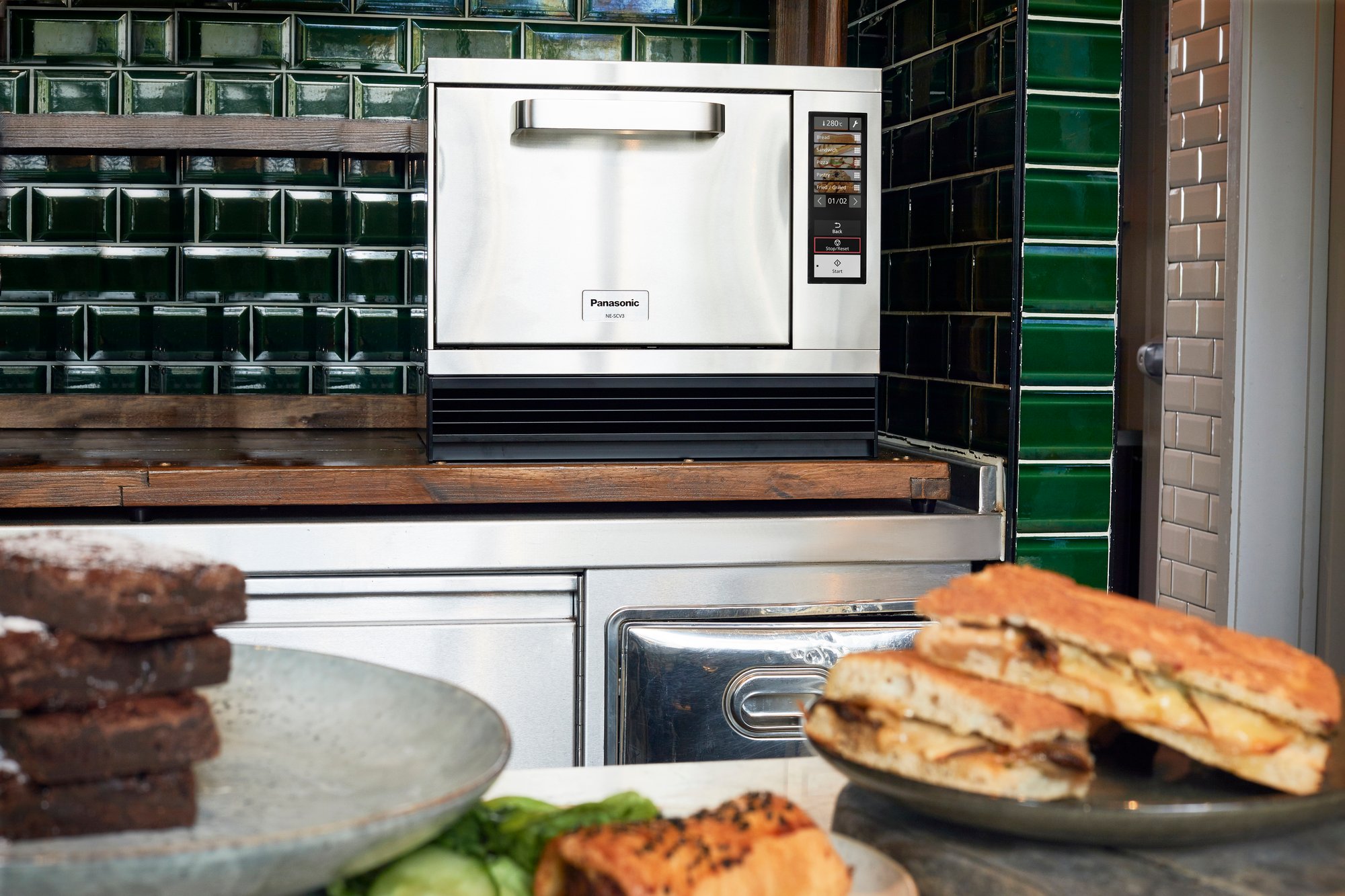 Panasonic NE-SCV3N high speed oven on the countertop with foods display in the forefront on another countertop.