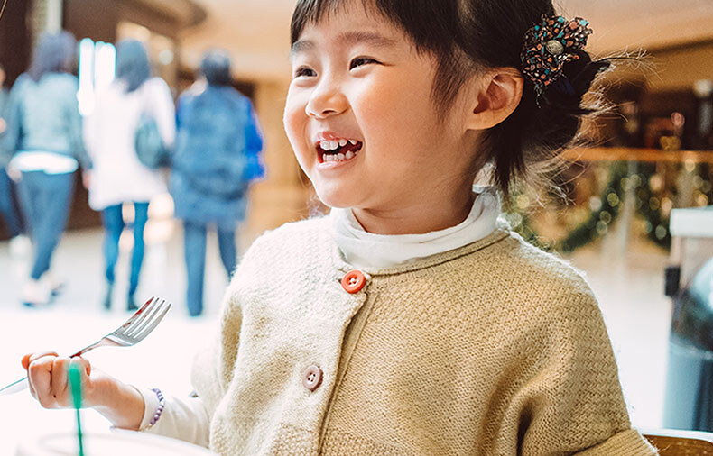 Young girl sitting in a food court holding a fork and smiling 