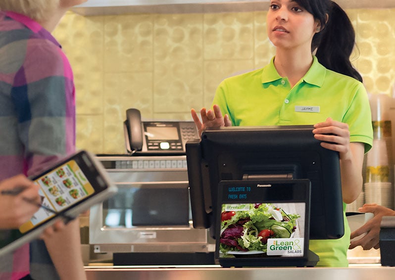 Woman standing behind POS machine, taking customer's order, while another customer orders using their phone