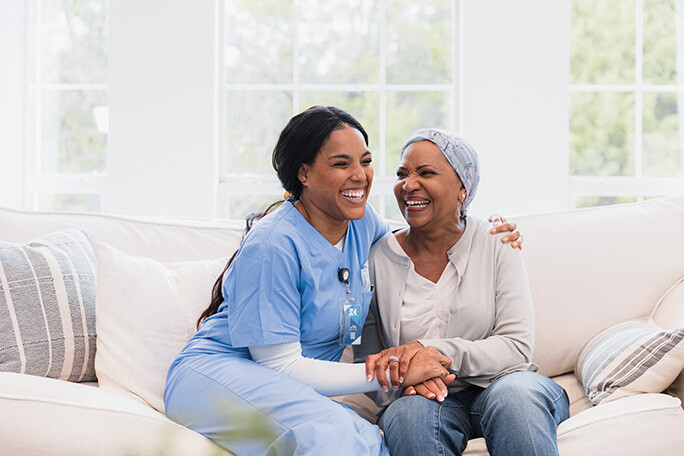 A nurse sits on a couch with her arm around a patient