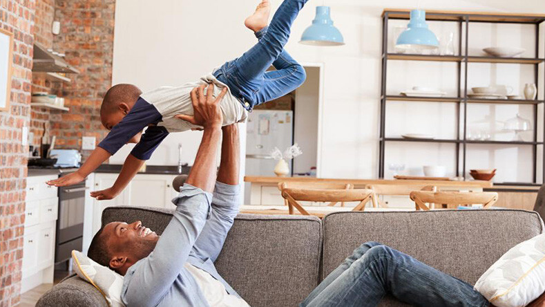A man holds his child in the air above him while laying on a couch