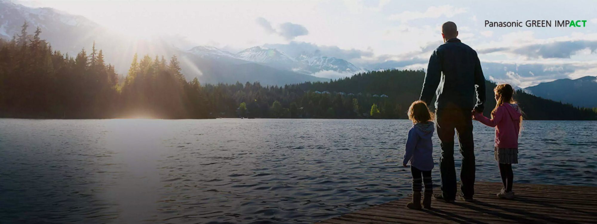 A father holds the hands of his two young children on a dock overlooking a lake