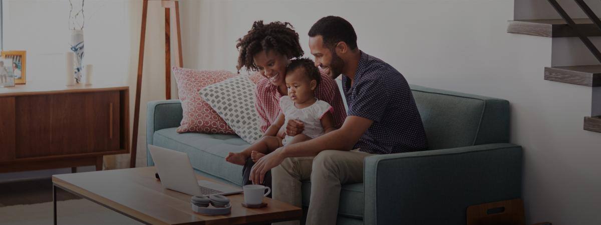 A smiling couple and their baby sit on a couch looking at a laptop