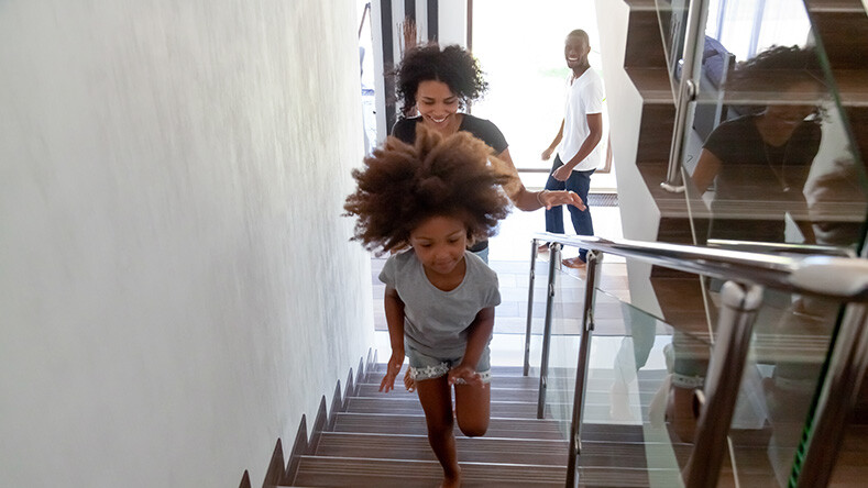 A smiling child runs up the stairs of her home as her mother walks behind her
