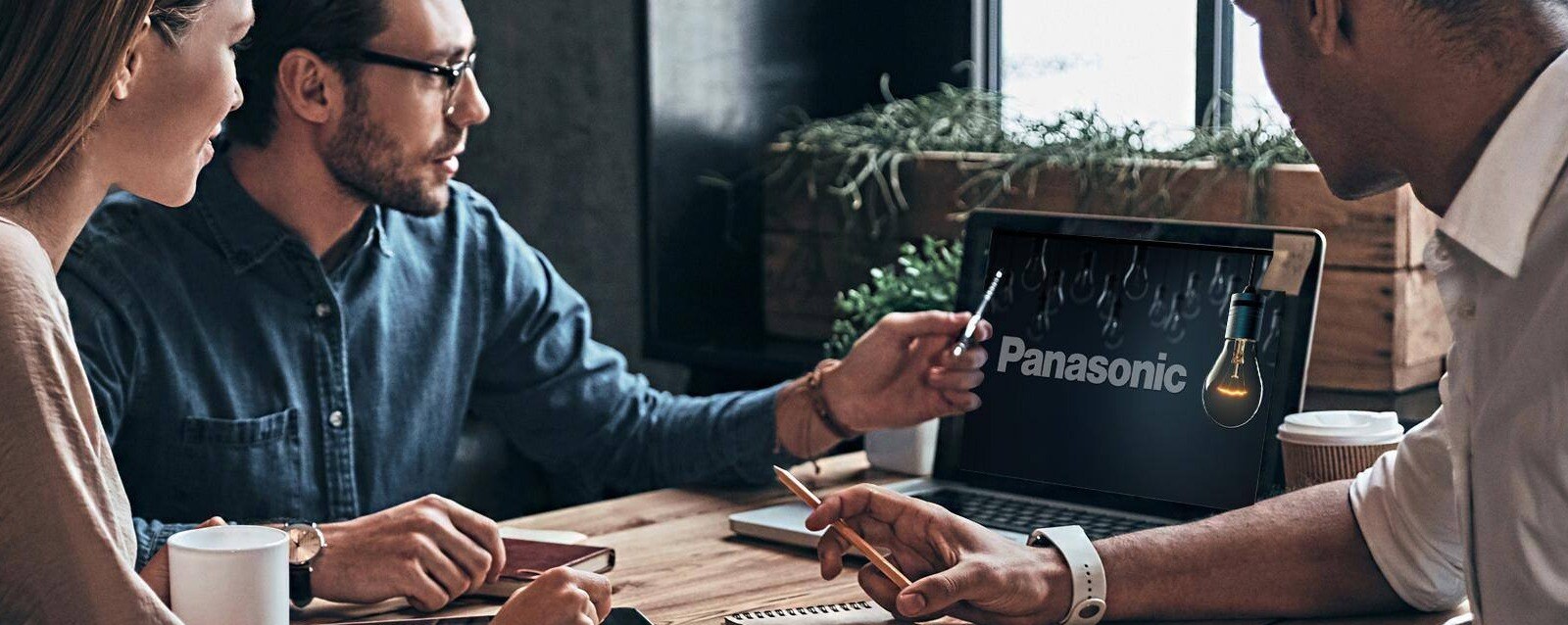 Three business people sitting around table looking at laptop screen that has "Panasonic" displayed on it