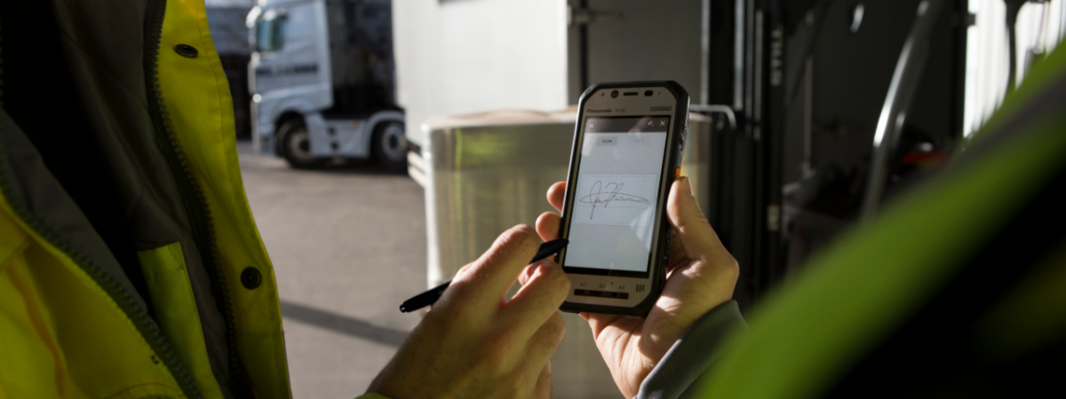 A hand holding a stylus leaves a signature on a phone screen and transport trucks sit in the background