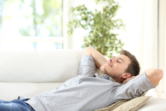 Man laying on a couch having a nap with arms crossed behind head havin