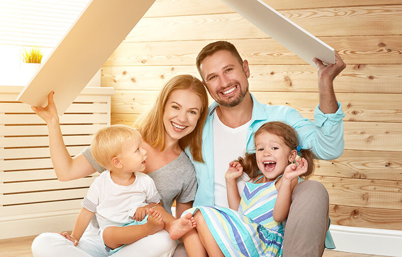 Man and woman sitting with young children in their laps, holding pieces of wood over their head in the shape of a house roof