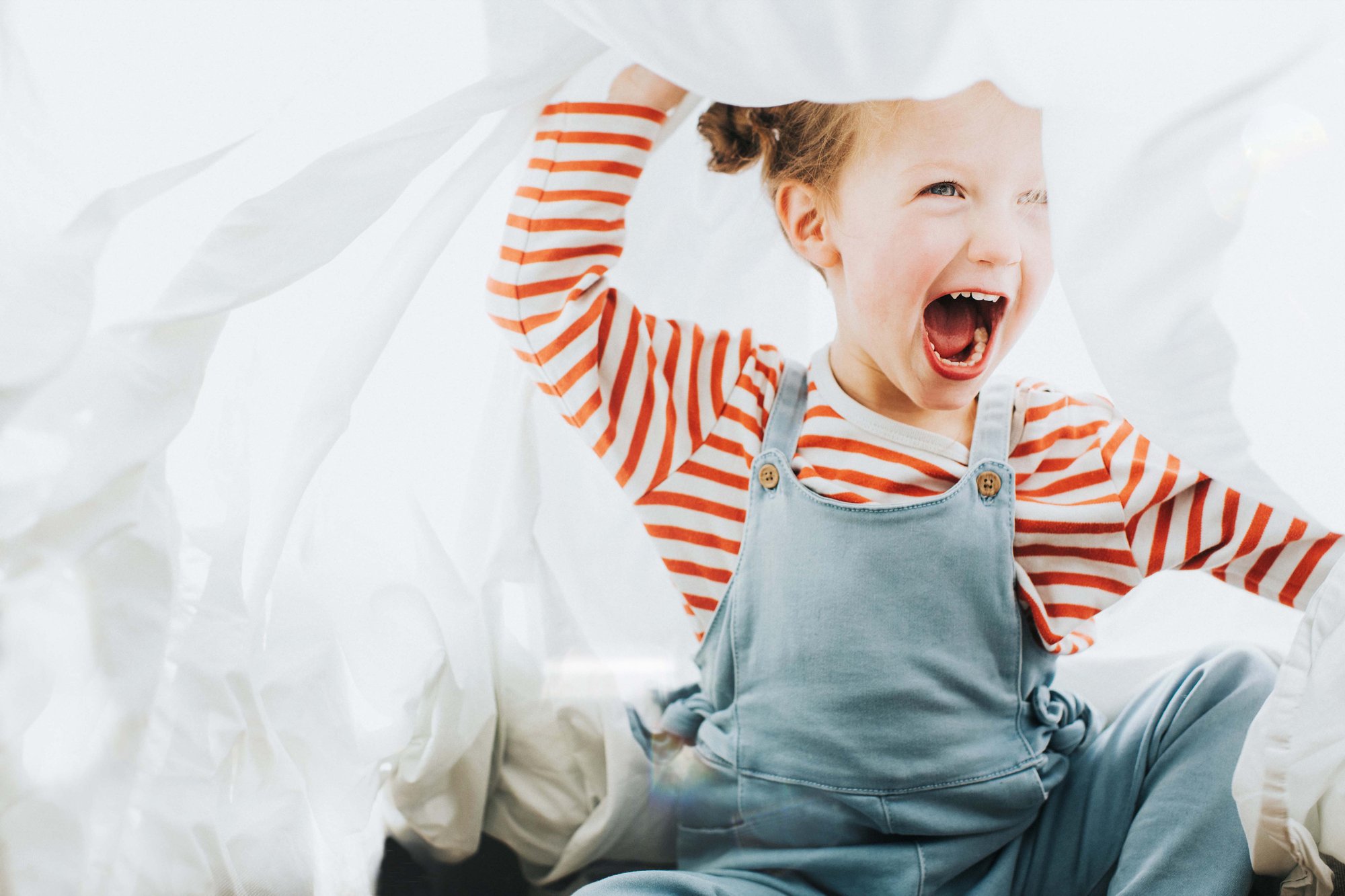 Little girl in blue overalls and red and white striped shirt laughing while playing under white sheets