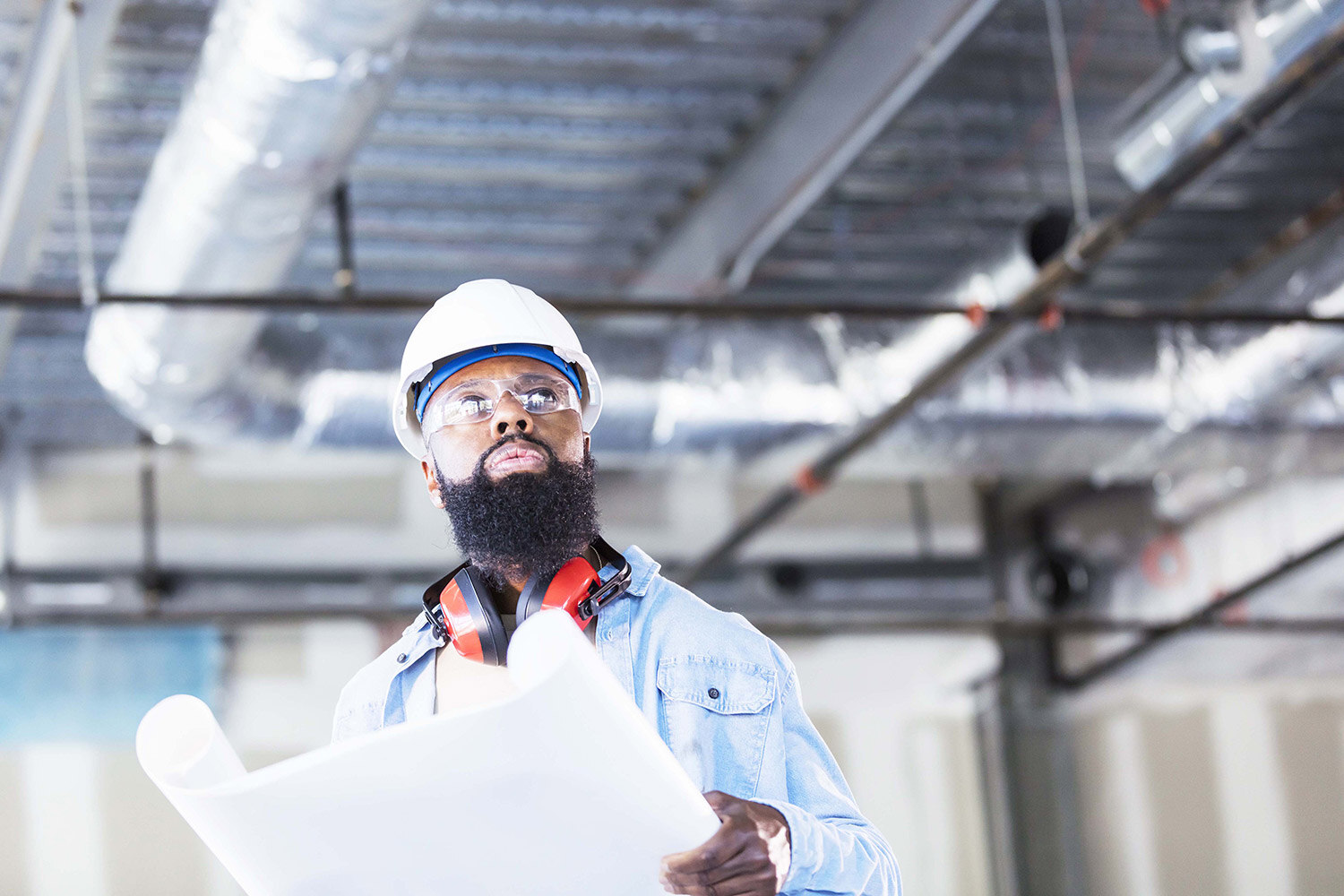 Man with white hardhat and safety glasses holding blueprint looking at ceiling