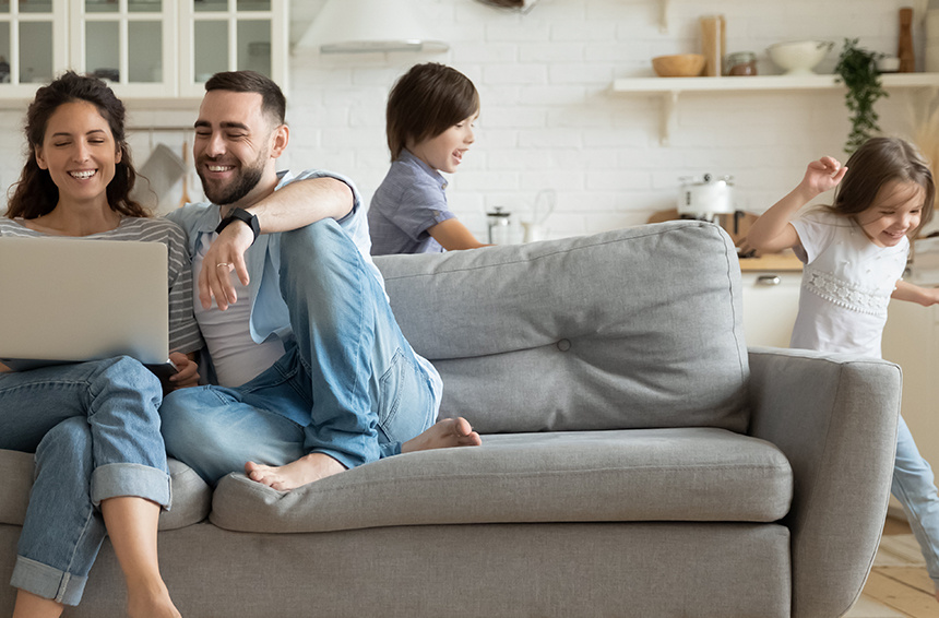 A smiling man and woman sitting on couch looking at laptop while children run around in background