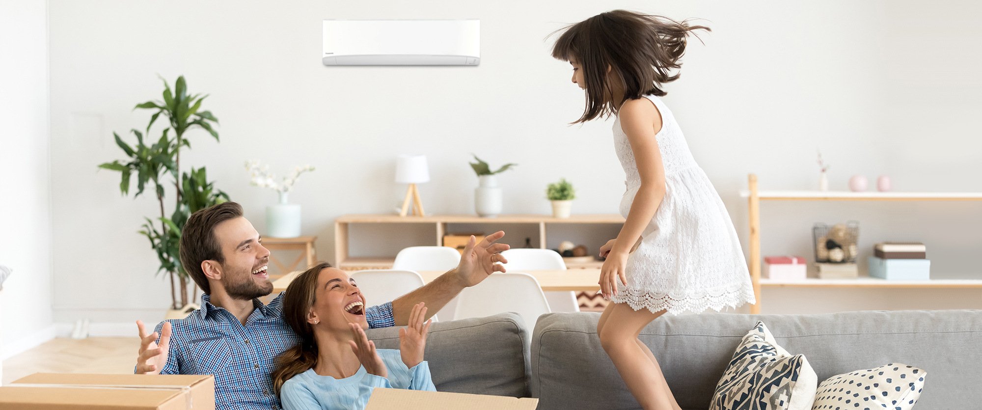 Couple sitting on couch with young girl jumping onto couch and air conditioner mounted on wall behind them