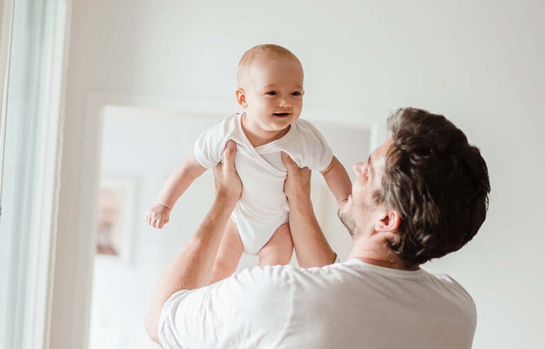 Man in white shirt holding baby in the air of brightly lit room