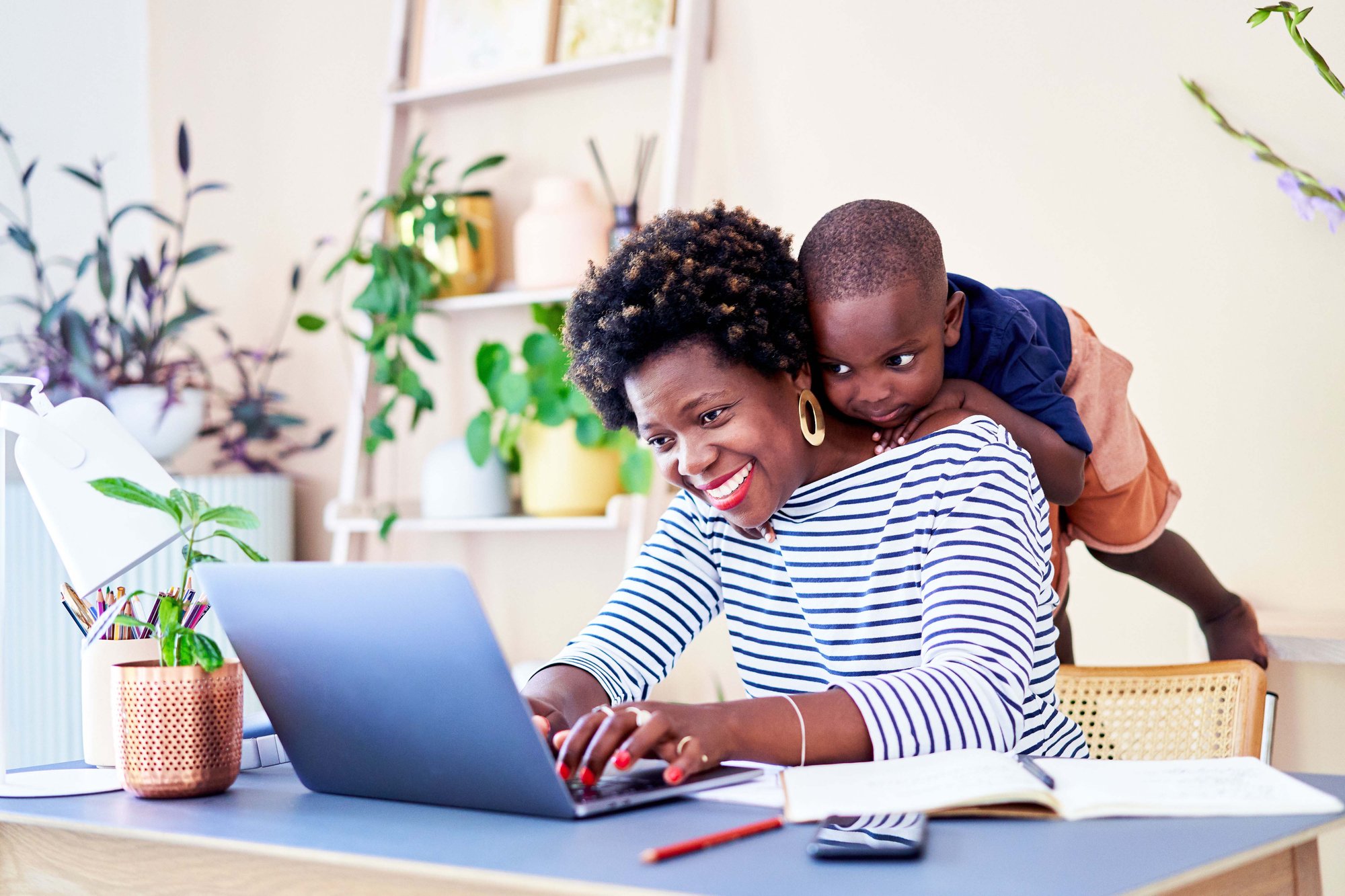 Woman sitting at computer with her child standing on the edge of her chair and leaning on her back to look at the screen