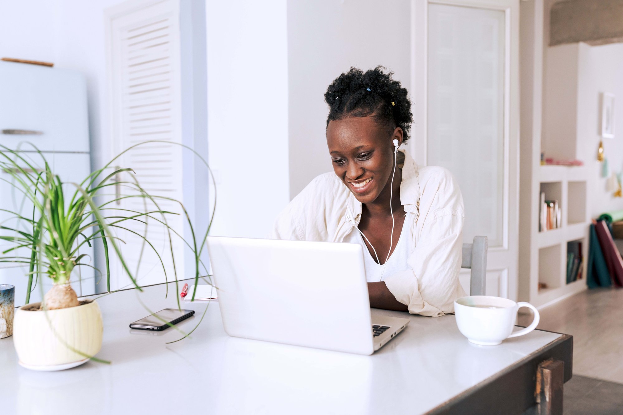 Woman wearing wired earbuds while smiling and working on laptop next to a coffee mug in a brightly lit room