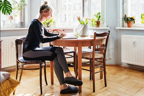 Woman sitting at wooden dining table working on laptop with sun shining through windows behind her