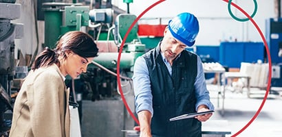 Woman and man staring at blueprints in a factory setting. The man has a blue hard hat on and a tablet in one hand while pointing at the blueprints