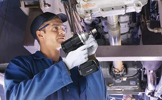 Man in mechanics suit wearing white gloves, blue hat and safety glasses, holding an assembly tool underneath of a car