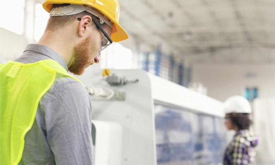 Man in yellow hard hat and high visibility vest looking at clipboard while woman in white hardhat watches C&C machine operate