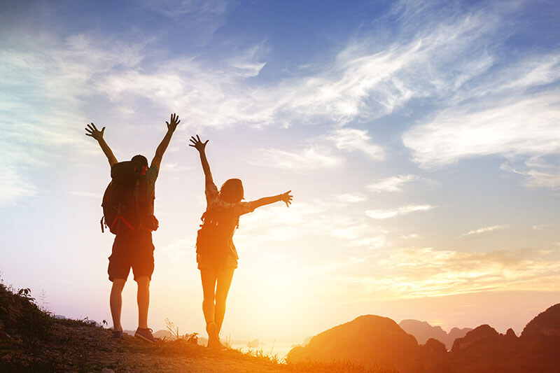Two hikers throw their hands up in the air facing a bright sunset