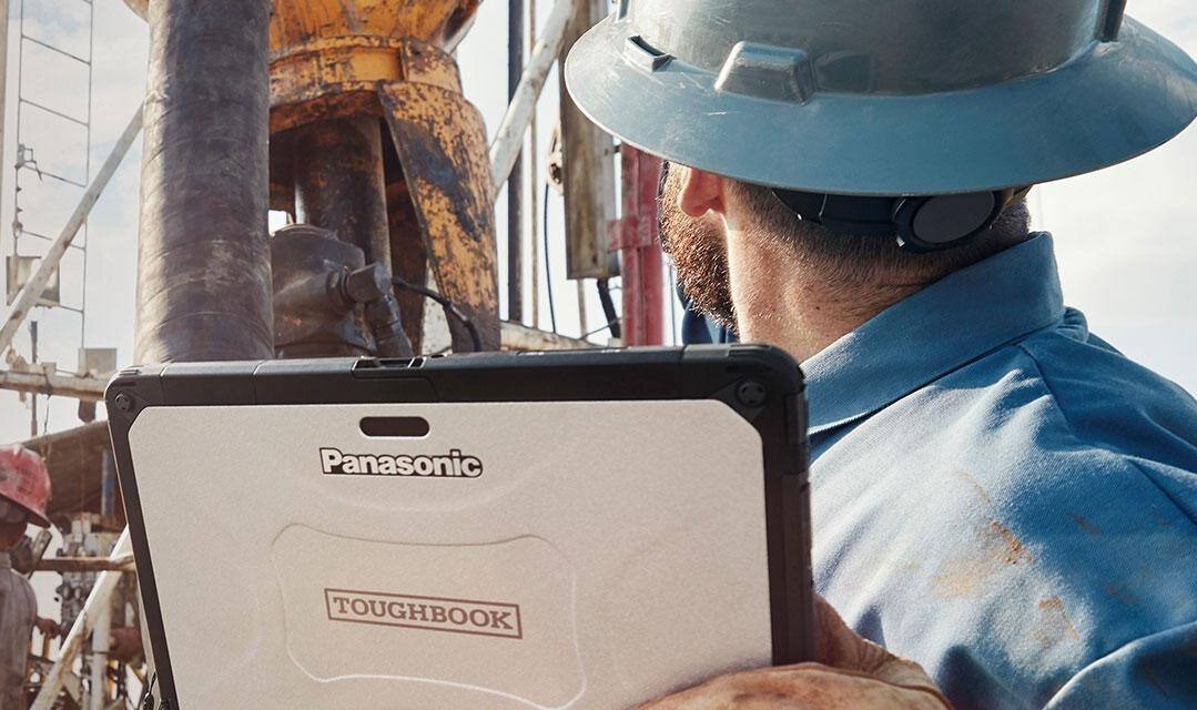 Man wearing hard hat holding a Panasonic Toughbook while looking at oil rig