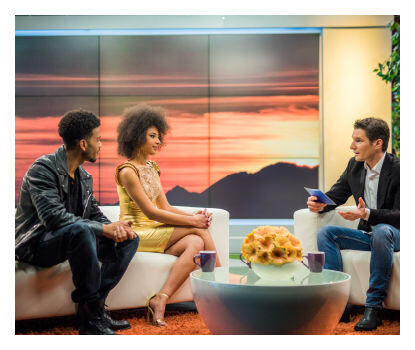 Trois personnes dans un studio de télévision avec toile de fond du coucher du soleil.