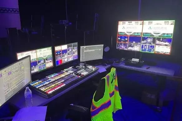 Broadcast control desk with multiple screens and equipment in a dark room.